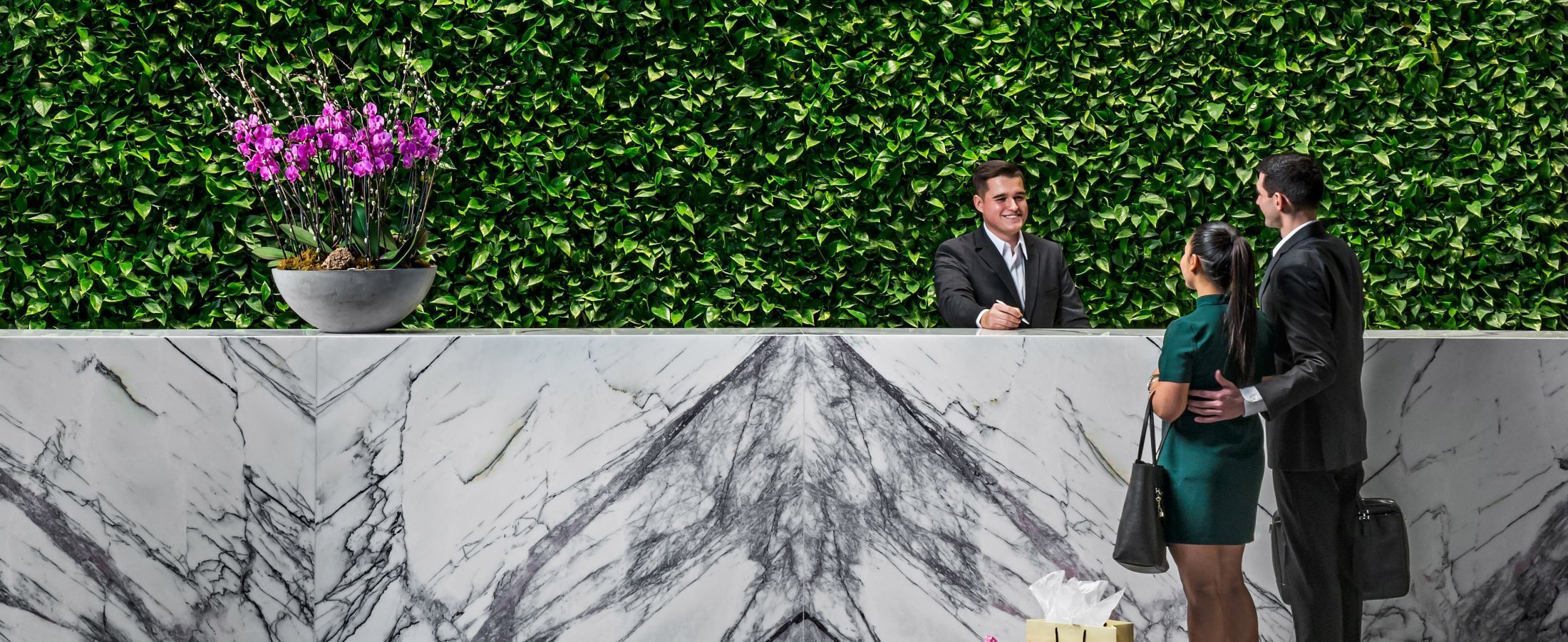 A front desk agent welcoming two guests at the large marble front desk with a green living wall behind it.